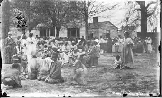 Historic black-and-white photograph showing a large gathering of African American men, women, and children outdoors, seated and standing in a circle as a woman speaks or leads from the right foreground. The group is dressed in post–Civil War–era clothing, with wooden buildings and bare trees in the background, suggesting a community meeting or church gathering in the Reconstruction South.
