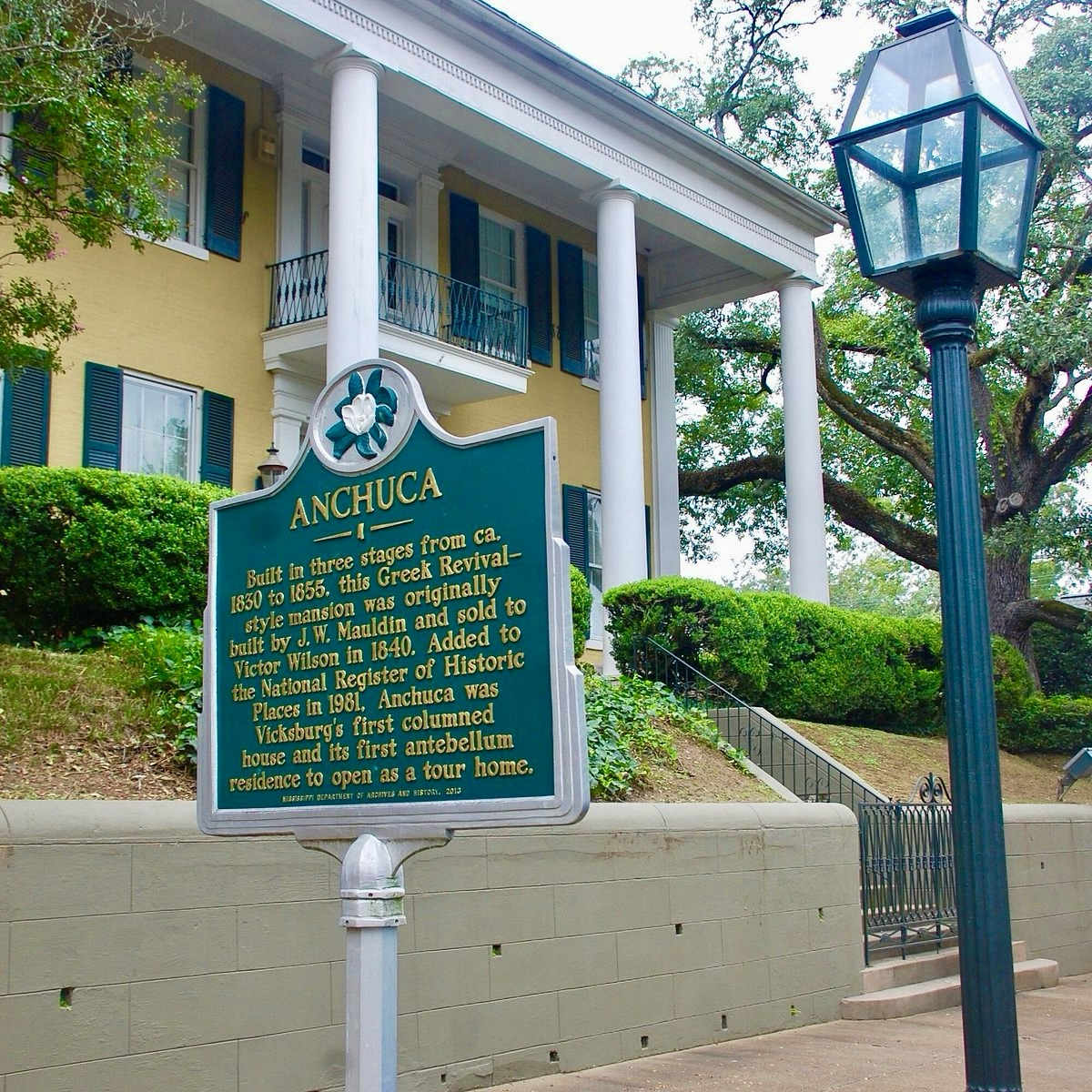 Color photograph of Anchuca, a Greek Revival–style mansion in Vicksburg, Mississippi. A historical marker in the foreground describes the home’s construction between 1830 and 1855 and its designation on the National Register of Historic Places. The yellow house with white columns and green shutters stands behind manicured shrubs and an old-fashioned streetlamp.