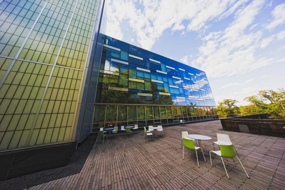 A building snakes in from the right most what we can see is a glass facade reflecting green forests and blue skies,  around it is an outdoor patio with occasional tables and chairs