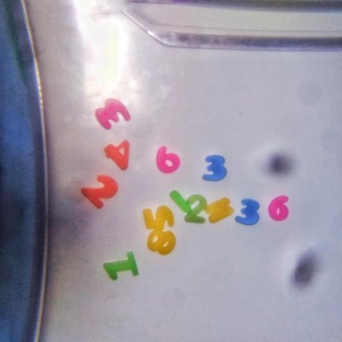 Una fotografía de un conjunto de números pegados a una puerta de heladera o refrigerador de color blanco . Hay siete números de colores brillantes colocados en un círculo o forma aleatoria. Los números son los siguientes: 1 (verde), 2 (azul), 3 (azul), 4 (rojo), 5 (amarillo), 6 (amarillo) y 6 (rojo). Los números parecen ser calcomanías o pegatinas de vinilo. Hay manchas oscuras de suciedad de la lente que esta manchando la imagen  alrededor de los números.