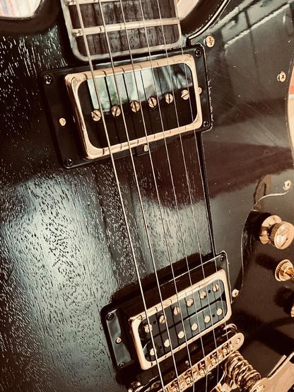 Eine schwarze E-Gitarre, Detailaufnahme des Korpusses, mit goldener Hardware. Die Gitarre ist schwarz-glänzend, die Holzstruktur aber deutlich sichtbar.
A black electric guitar. Close-up shot of the body. Golden hardware. The guitar is glossy black, but the wood structure is clearly visible.