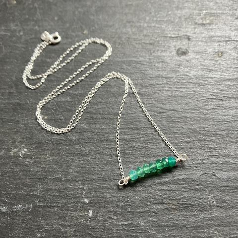 A photo of a delicate, handmade necklace featuring a row of seven green Chalcedony beads hanging from a silver chain & c-clasp on a dark grey slate background.