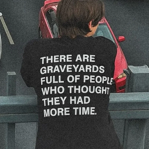 Person leaning over a railing in a parking lot wearing a black T-shirt with white block text: "THERE ARE GRAVEYARDS FULL OF PEOPLE WHO THOUGHT THEY HAD MORE TIME."