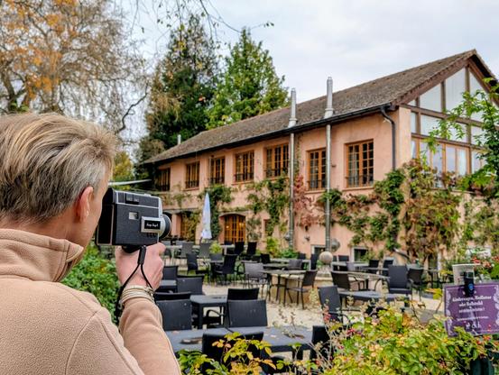 A person films a rustic, ivy-covered building with large windows and outdoor seating, using a vintage-style video camera. The scene includes tables and chairs arranged in a garden setting, with a sign in the foreground about accessibility for strollers and wheelchairs.