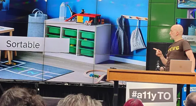 Adrian at the #a11yTO podium pointing to a slide titled "Sortable" that shows a white box with 3 rows and columns of green plastic drawers. Adrian is wearing a brown T-shirt with yellow letters W C A G, each letter separated by asterisks, like in the title of the old TV show MASH.