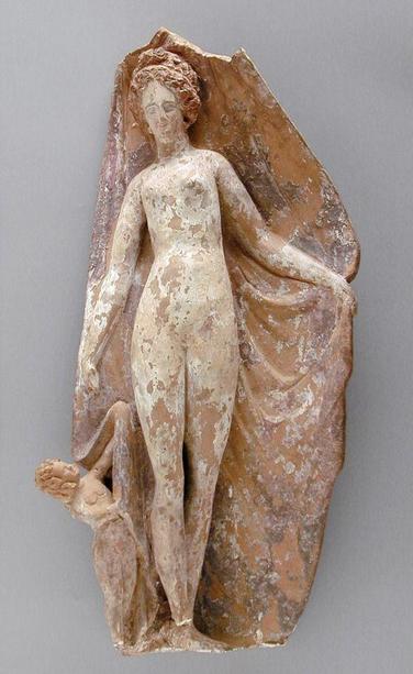 Nude Aphrodite terracotta figurine with paint residue showing that her skin is pale and her hair a reddish brown. Eros holds the cloth of a cloak or himation that falls behind her, looking up at her adoringly. His hair was painted blond.