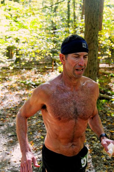 Man running with a black cap but no shirt in a forest with trees lit up yellow with sunlight and fall color