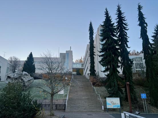 A view of the white blocky buildings of Ylistö campus. In the center, stairs go up to the buildings. On the right, there are thin spruces. On the left, there's an abstract sculpture.