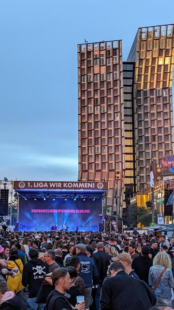 Bildbeschreibung von KI:
Das Foto zeigt die Tanzenden Türme in Hamburg, zwei markante, leicht gegeneinander geneigte Hochhäuser aus Glas und Metall, die in der Abenddämmerung in warmen Tönen leuchten. Im Vordergrund steht eine große Bühne auf dem Spielbudenplatz mit der Aufschrift 1. Liga wir kommen. Auf der Bühne sind mehrere Musiker zu sehen, über ihnen leuchtet in pinker Schrift der Name Dangerfabrique. Davor hat sich eine dichte Menschenmenge versammelt, die ausgelassen feiert. Viele tragen schwarze Kleidung, einige mit dem Totenkopfzeichen des FC St. Pauli. Manche halten Getränke in der Hand, andere tanzen oder reden miteinander. Im Hintergrund erstrahlt die Fassade der Türme im Licht des Sonnenuntergangs, während über der Szene der Himmel in hellem Blau erscheint. Die Stimmung ist fröhlich und festlich, es wirkt wie ein besonderer Moment des Stolzes und der Gemeinschaft.
