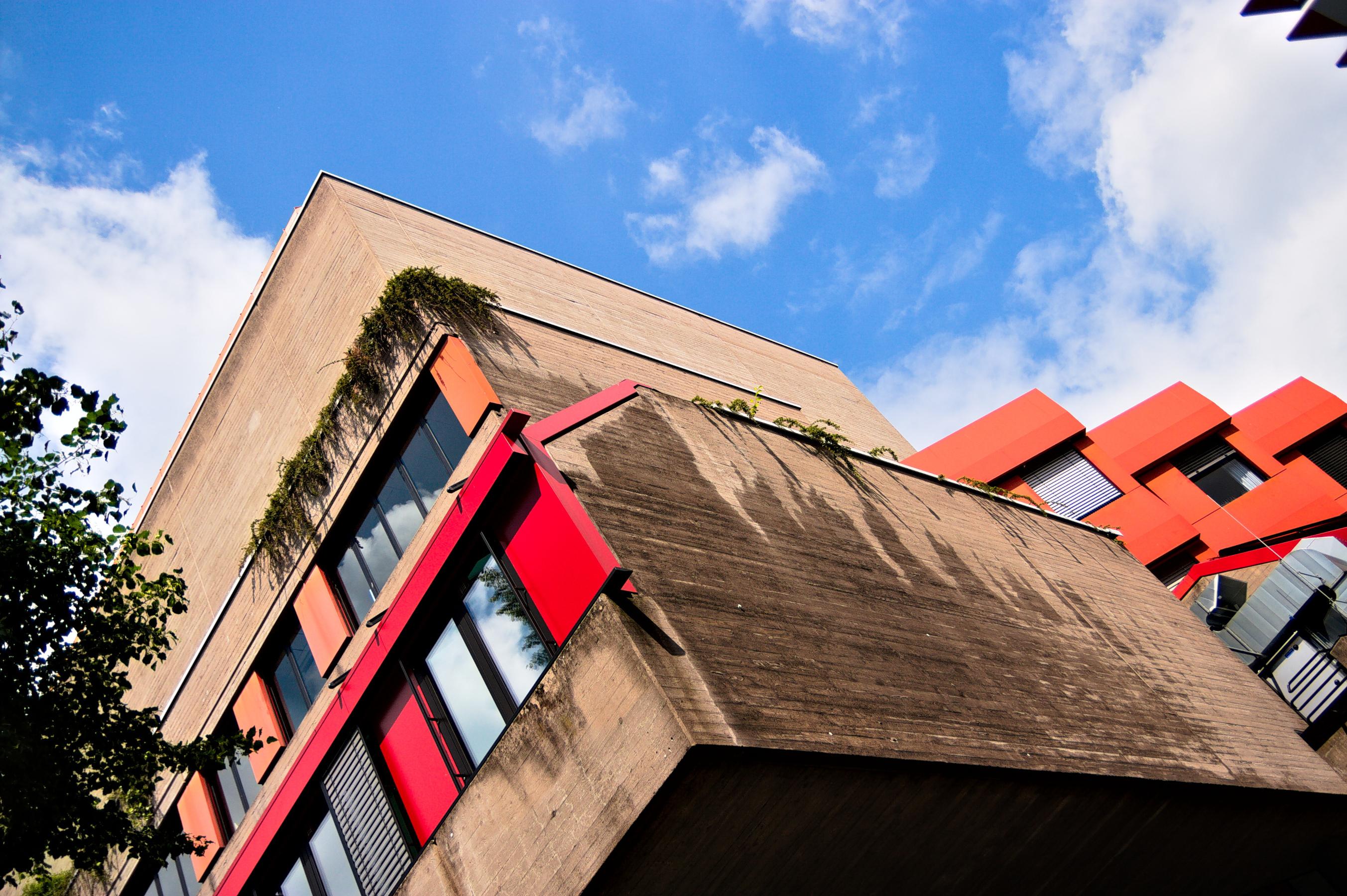 Das Bild zeigt das Gebäude der Musikhochschule in Köln. Das Gebäude ist im Stil des Brutalismus, also mit rohem Beton, gebaut. Fassenelemente in hellrot setzen sich davon ab. Die Kamera wurde bei der Aufnahme verkippt, so dass die Linien des Gebäudes nach links weg kippen.