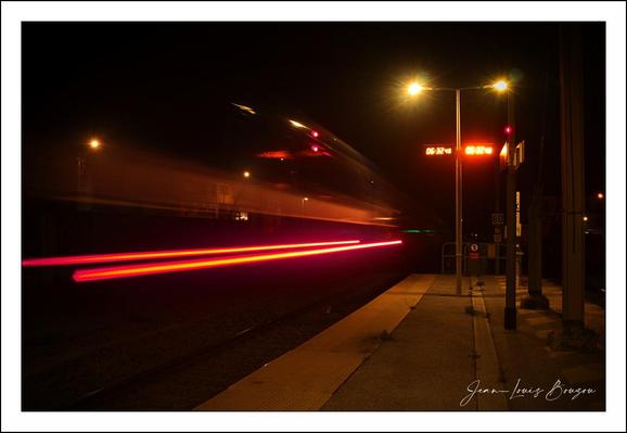 This image captures a night scene at a train station with a remarkable sense of motion and light. The focal point is a train speeding through the station, rendered almost ghostly and blurred due to its swift movement and the long exposure of the camera. The train's lights streak horizontally across the frame in intense red and white lines, creating a dynamic contrast against the deep black night sky and the dark silhouettes of the surrounding environment.
The platform and the lamppost emit a warm yellowish glow that softly illuminates a small section of the station, lending a quiet, almost lonely atmosphere to the setting. The light also reflects off the platform surface, enhancing the depth and perspective. This combination of the stationary, softly lit platform and the rushing blur of the train visually conveys a theme of transition — the stillness of waiting versus the relentless passage of movement.
In terms of symbolic or cultural meanings, trains often represent journeys, both physical and metaphorical, and the contrast between the static platform and the fleeting train can evoke reflections on time, change, and fleeting moments. The stark lighting and night backdrop might suggest themes of mystery or the unknown paths ahead.
Artistically, the photograph is a powerful use of exposure and light, capturing not just a moment but a temporal flow. The red streaks of light add a dramatic flair that heightens the emotional impact of the scene.
