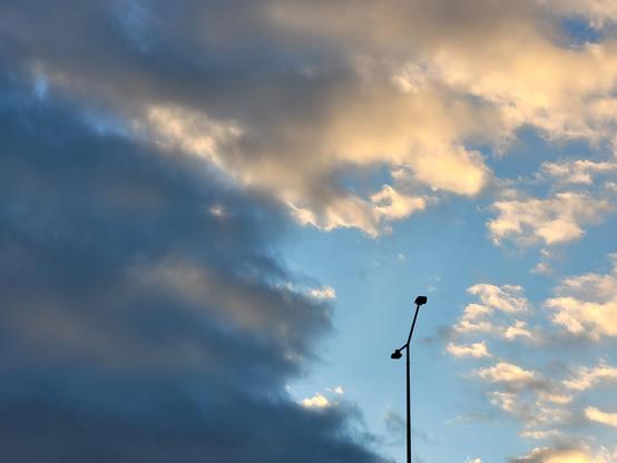 A photo of the sky, a single, thin streetlight shooting up from the bottom right, dark against the lighter background, no trees or street visible. The left side of the sky is covered by dark grey clouds, their tattered edges leave room on the right for lighter altocumulus clouds to shine in the autumn evening sun. The streetlight is framed almost perfectly by a patch of blue sky between the clouds.
