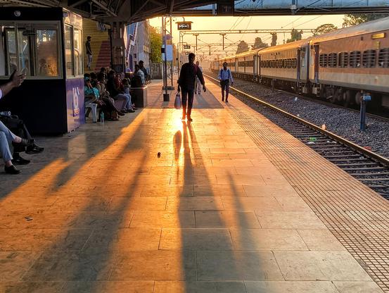 Long shadows stretch across a train station platform as the setting sun casts a golden glow. A train is visible on the tracks to the right, and people are waiting on the platform, some seated, others walking. The warm light highlights the figures and the texture of the platform.