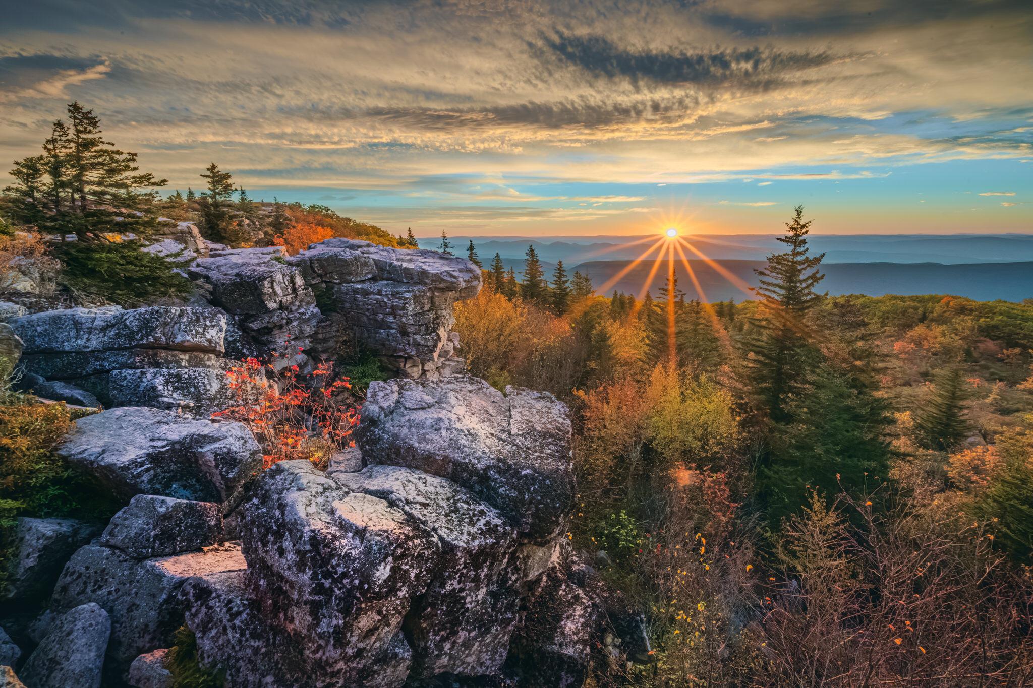 A sunrise over a valley with textured rocks in the lower left of the frame, fall colored trees towards the right leading to the midground with layers of the valley beyond. The sun has the sunburst effect as it crosses the horizon with a light layer of clouds overhead.