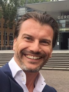 Een lachende Vindent, donkere haren en snor en baardje. Blauw jasje, wit overhemd.