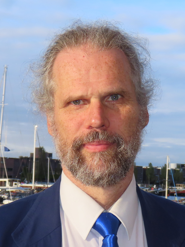 Foto van André; grijze, beetje wilde haren, snor en baardje, blauw jasje over een wit overhemd met een felblauwe stropdas. Op de achtergrond een haven met zeilboten.