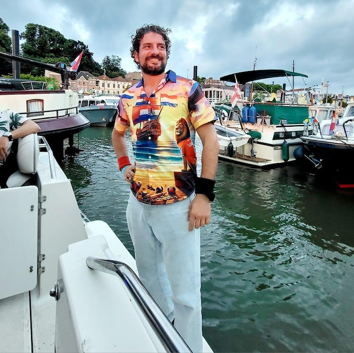 Foto van een lachende Roberto op de rand van een boot in een haven. Donkere haren, snor en baardje, bontgekleurd overhemd (met o.a. prints van een leeuw en een bootje met Nederlandse vlaggen) op lichte broek. Bootjes op de achtergrond.