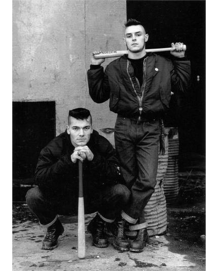 Julien Terzics and his friend Jeff. They are dressed in a fairly typical 80s punk/hardcore aesthetic with cuffed jeans, combat boots, and light jackets. Julien is squatting resting his chin on a bat. Jeff is standing casually holding a bat behind his neck across his shoulders.