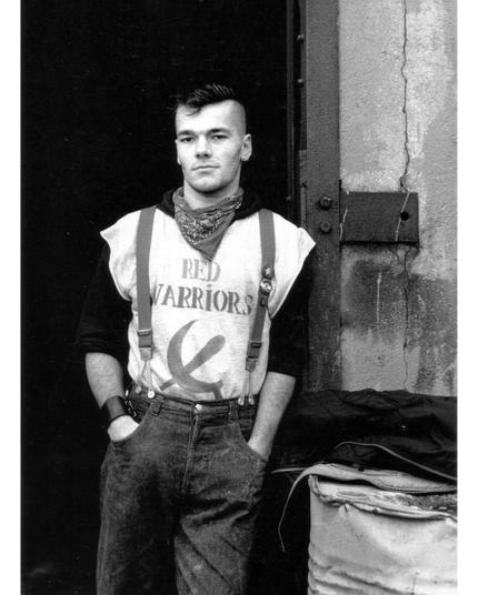 Julen leaning against a door jamb. He's wearing a shirt that says "red warriors" with a hammer sickle on it, suspenders and a bandana as a scarf.