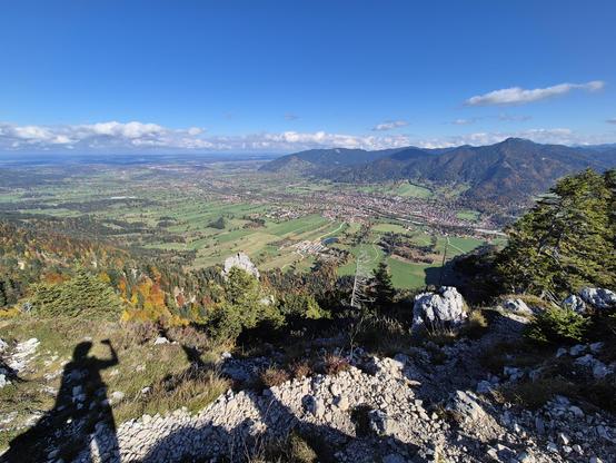 A breathtaking panoramic view from a mountaintop, showcasing a vast and vibrant valley below. The landscape is a patchwork of lush green fields, clusters of colorful autumn trees, and quaint villages nestled among rolling hills. Majestic mountain ranges stretch into the distance under a clear blue sky, creating a sense of awe and tranquility. The rocky foreground adds texture and depth, emphasizing the grandeur of the scene.