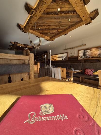 A cozy and inviting interior of a rustic restaurant or café, featuring warm wooden beams, benches, and tables. The foreground showcases a menu with the elegant inscription "Landerermühle," resting on a polished wooden table. The background reveals a charming seating area with plush cushions, framed pictures of scenic landscapes, and soft lighting, creating a welcoming and homely atmosphere perfect for relaxation and enjoyment.