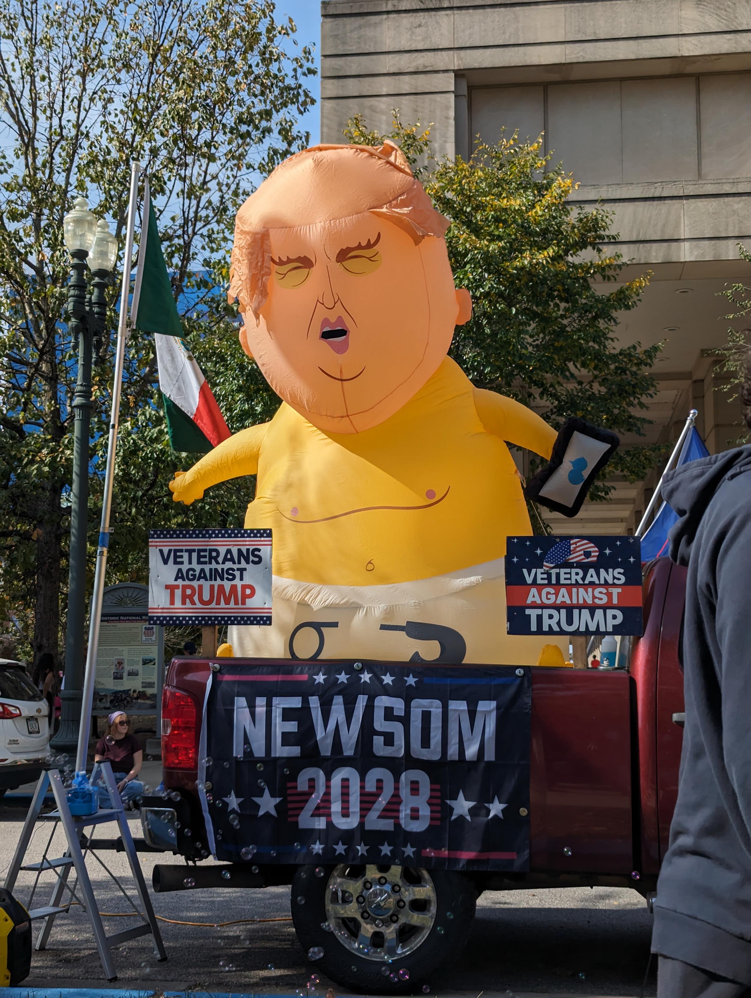 Giant baby balloon in truck bed with signs that say newsom 2028, veterans against trump