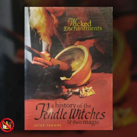 A photo of a large format softcover book, "Wicked Enchantments – a history of the Pendle Witches & their magic" by Joyce Froome, is inserted into a cropped and dimmed image of itself to frame the image.

The photographic cover depicts a woman in a robe, muddling with a large pestle and mortar, on a table covered with a red cloth. A black cat sits on the edge of the table sniffing at scraps of ingredients for the concoction.

An emoji crown in the lower left corner of the frame is superimposed with the red circle and slash emoji.