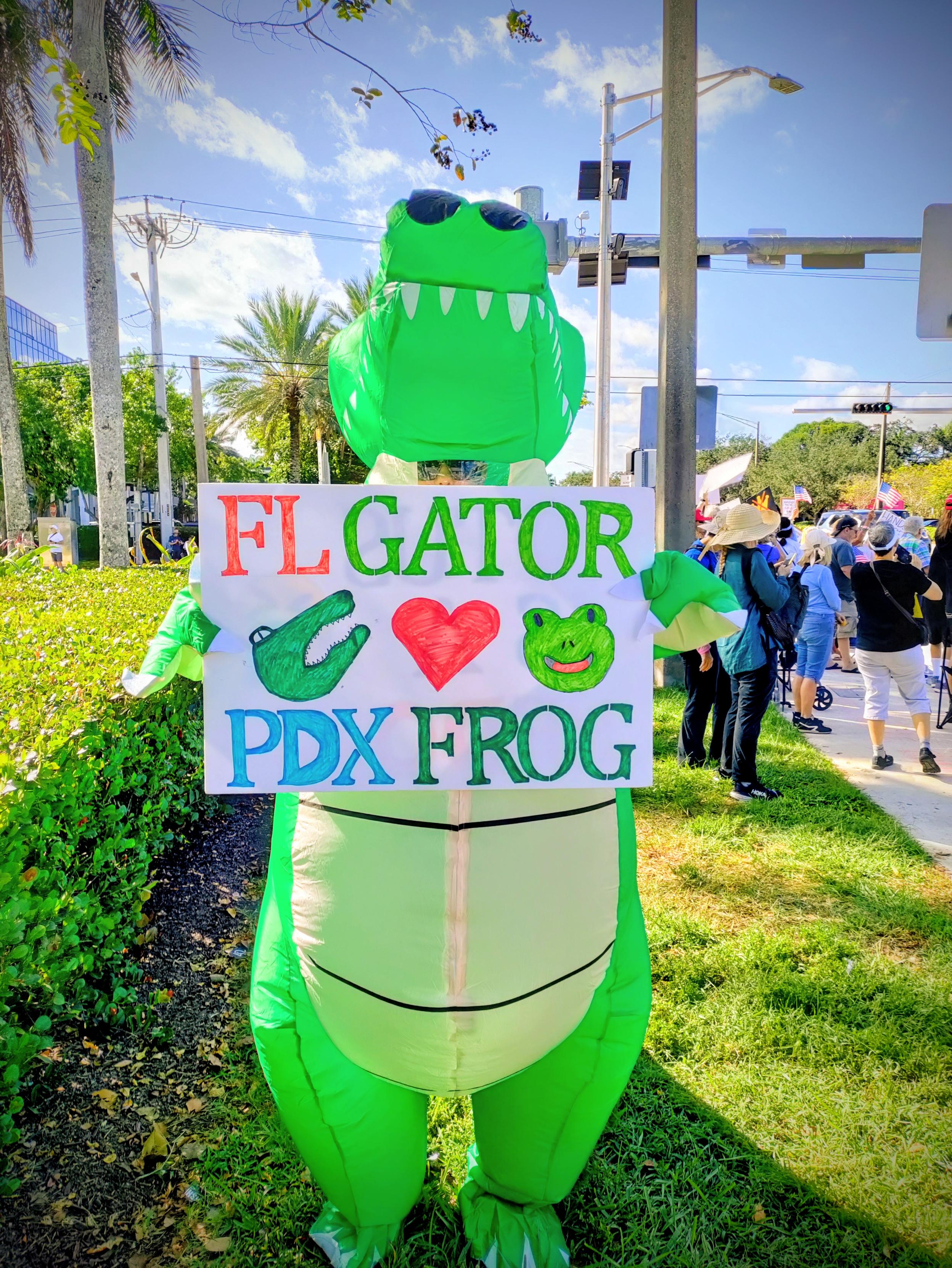 Woman at No Kings Protest  in inflatable gator costume holding a sign that states "FL Gator ❤️ Portland Frog".