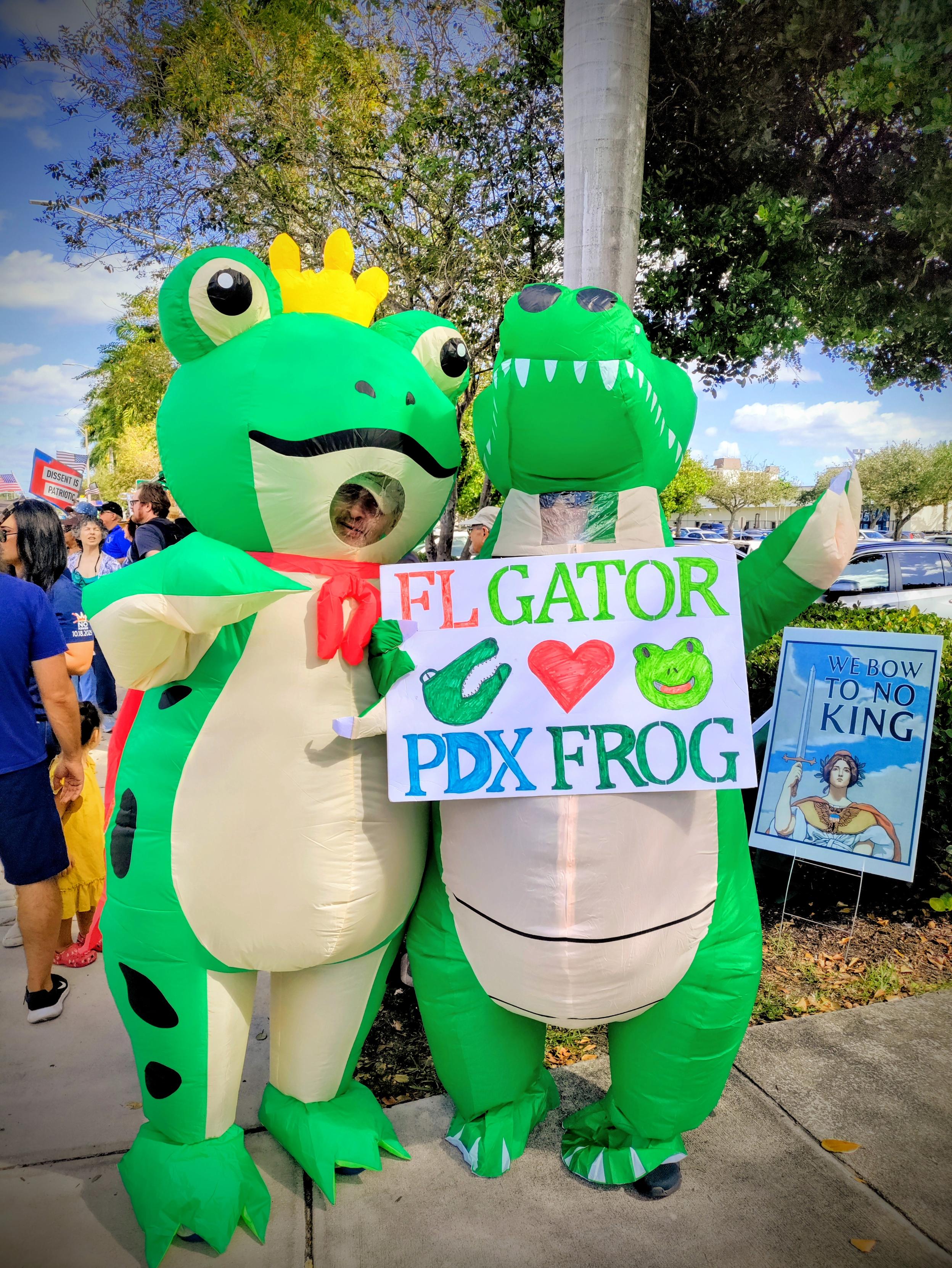 Man in the classic infaltable Portland frog costume next to lady in an inflatable gator costume holding a sign that reads "FL Gator ❤️ Portland Frog".