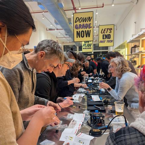 A crowded workshop table filled with people soldering and assembling small electronics during a hands-on repair event. Signs above read “DOWNGRADE NOW” and “LET’S END FAST TECH.”