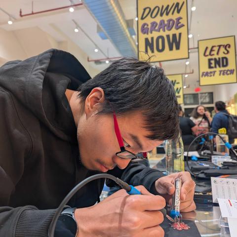 A focused participant uses an iFixit soldering iron to assemble a small LED circuit board at a community repair event, with bright yellow signs in the background promoting sustainable tech.
