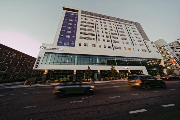 Many-storied building with a blue stripe to the left looms over a road with several cars blurred by motion