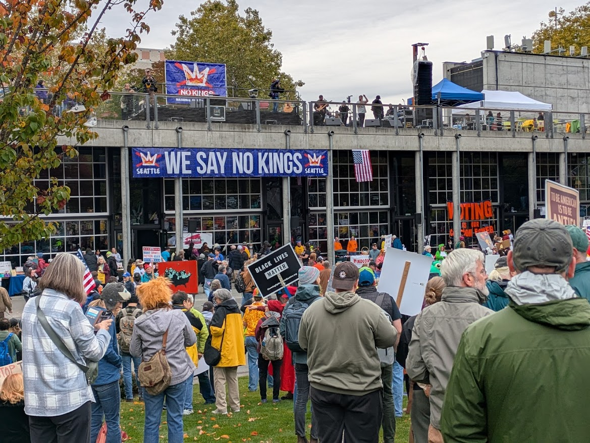 People gathering at Fisher Pavilion in the Seattle Center with their anti-fascist, pro-democracy signs; speakers and band are on top of the exhibition building and look a little small and remote, but could be heard loud and clear.