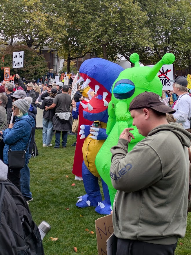 Playful costumes at the rally included aliens, frogs, more frogs, and a variety of colorful, grinning dinosaurs. A little larger than life here are a green alien and a blue-white-red dinosaur.