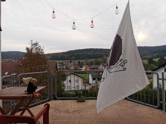Blick über einen Balkon mit einem kleinen, bepflanzten grünen Blumenkasten auf dem Boden in der Mitte vor dem Geländer und einem dunkelgrünen bepflanzten Blumenkasten links am Geländer. Links steht eine rote Bank und ein Holztisch mit einem Plüschraben mit wollweißer Mütze darauf, rechts hängt eine große Korsikaflagge. Oben hängt eine Kordel mit vier Solarleuchten quer. Im Hintergrund ein Dorf. Durch die geschlossene Wolkendecke ist rechts der Flagge die Sonne zu erahnen.
