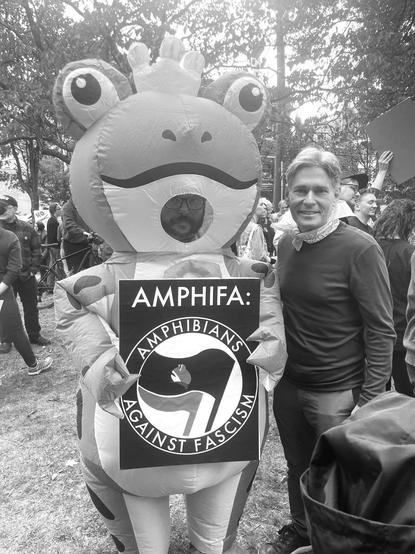 Demo-Teilnehmer in Froschkostüm hält Schild vor sich mit dem Antifa-Logo plus Schriftzug „AMPHIFA - AMPHIBIANS AGAINST FASCISM“