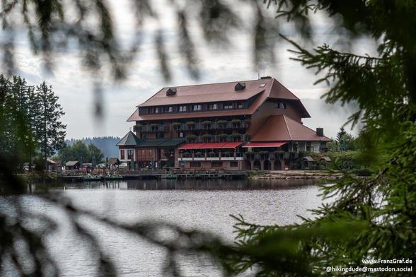 Framed by delicate pine needles, a charming lakeside lodge emerges—its rustic red roof and wooden balconies blending seamlessly with the surrounding forest. The multi-story building, with its warm brown tones and flower-adorned railings, exudes alpine coziness. Below, a calm lake mirrors the muted overcast sky, its surface undisturbed except for a few small ripples. The lodge’s lower level extends almost to the water’s edge, inviting guests to step outside and enjoy the tranquil setting. In the…