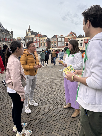 Twee D66-vrijwilligers praten op een stadsplein met een man en vrouw. Op de achtergrond staan kleurrijke oude huizen en marktkramen. De sfeer is open en betrokken; er wordt rustig geluisterd en informatie gedeeld.