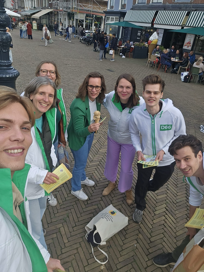 Zeven vrijwilligers van D66 staan lachend bij elkaar op een plein met historische panden. Ze dragen groene en witte jassen met het D66-logo en houden flyers vast. In het midden houdt een vrouw een ijsje vast. De sfeer is vrolijk en energiek.