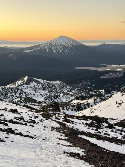 A view looking down the climbing route of South Sister. A prominent volcano, Mount Bachelor, stands in the distance. The popular ski destination has white snow covering the top of the mountain. White snow covers much of the climbing route below. An orange sunrise fills the sky above. Photo taken while climbing South Sister on October 18, 2025, in the Three Sisters Wilderness in central Oregon, U.S.