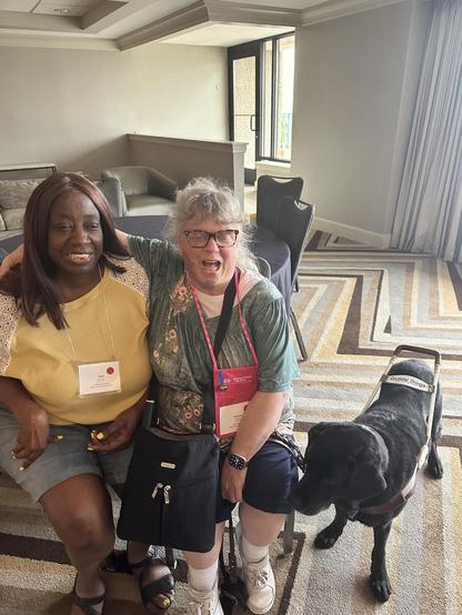 Picture of Toni Burrell (left) and Danette Dixon (right) with her guide dog Mr. Mayor from Guide Dogs for the Blind. They sit in a hotel meeting room at the 2024 ACB Convention. Danette has her arm around Toni’s shoulders, and they both smile at the camera. 