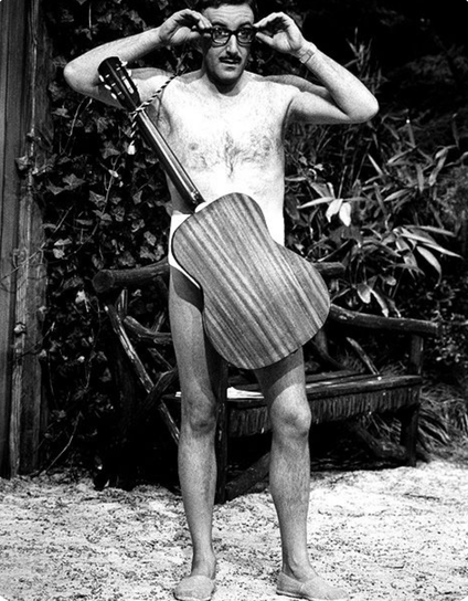 A black and white still from the film "A shot in the dark" which features a naked Peter Sellers visiting a naturist park. covering himself with a guitar