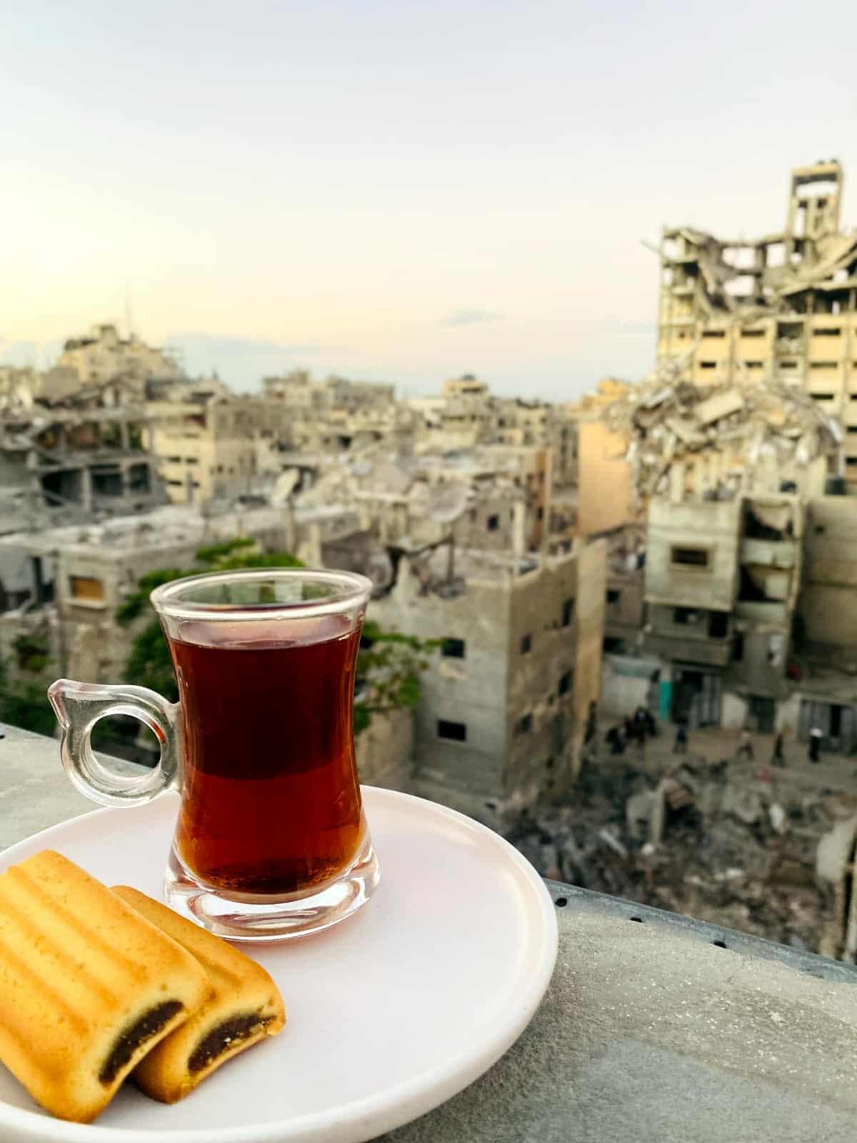 Picture of a white plate with two biscuits on it and a cup of tea. The point of view seems to indicate it's taken from a rooftop. The background is the blurred view of a city in rubble in the Gaza Stripe.