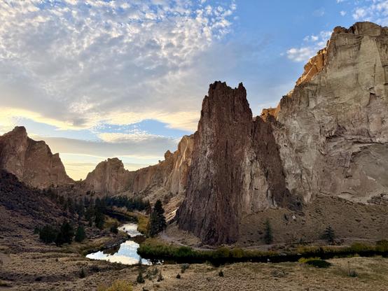A scenic view of towering rock formations at sunset, with a winding river reflecting the sky. The foreground features greenery and trees, while the background showcases dramatic cliffs under a cloudy sky.