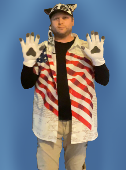 I'm standing, wearing the clothing I wore at No Kings Day 2: beige pants, black turtleneck, button-down shirt bearing the image of an American flag and the preamble of the United States Constitution, white gloves with paw prints on the palms, a cosplay hat with my border collie OC's face and a facemask with his muzzle.  I'm holding my hands up to show the beans.  The mask is hanging by the side of my face. Background removed, replaced by slightly muted blue.