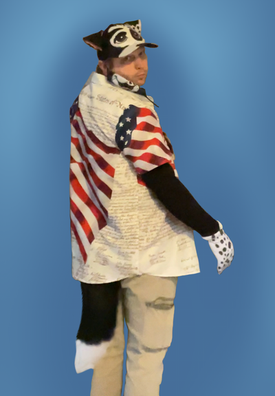 Same me, same outfit, but now turned to a quartering-away pose to show off the fluffy black-and-white tail that was also part of my costume.  The back of the right glove is visible, and it is covered with black spots, like my OC's right paw.  Same background as previous photo.