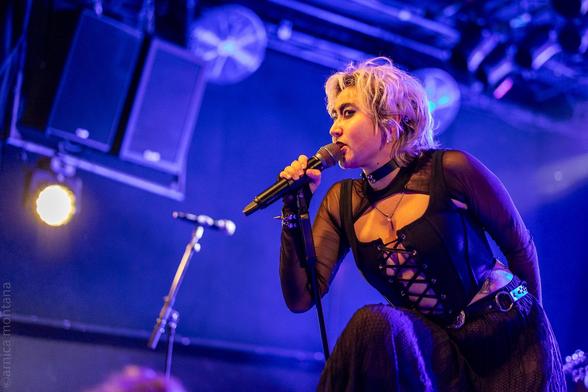 A performer on stage singing into a microphone, dressed in a black lace-up top with a sheer overlay. The background features stage lights and sound equipment, creating a vibrant concert atmosphere.