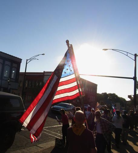 A street scene with people peacefully demonstrating. In the center of the picture, an American flag is illuminated by the sun setting behind it. 