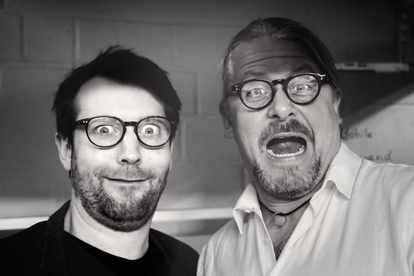 S/W-Foto von zwei Männern mit Brille und Bart, die Grimassen schneiden. 