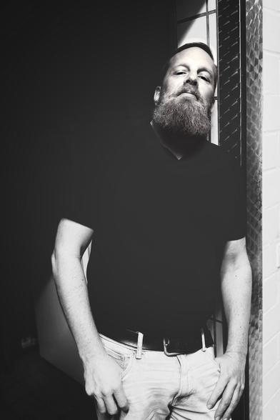 S/W-Bild eines Mannes mit kurzen Haaren und einem längeren getrimmten Bart in schwarzem Poloshirt, der lässig an einem Türrahmen steht. 